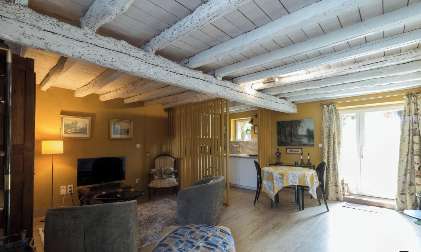 A room with a wooden ceiling, a television, a dining table with chairs, and a window with curtains.