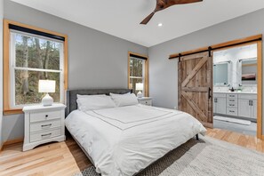 Ein Schlafzimmer mit einem Bett, Nachttischen, einer Kommode, einem großen Fenster und einer gleitenden Stallschranktür, die ins Bad führt.