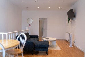 A modern living room with a sofa, dining table, chairs, a small coffee table, a flat-screen TV mounted on the wall, and a radiator. - Apartment Sleeps 4 With Wifi & Garden (Bolton)