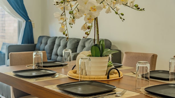 Una mesa de comedor dispuesta con platos, cubiertos y copas, con una planta en maceta y un jarrón de flores en el centro.