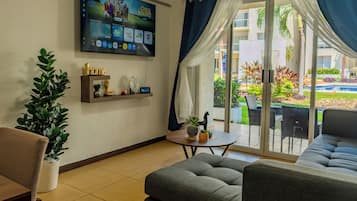 A modern living room with a flat-screen TV, a sofa, a coffee table, and a view of a garden through a sliding glass door.
