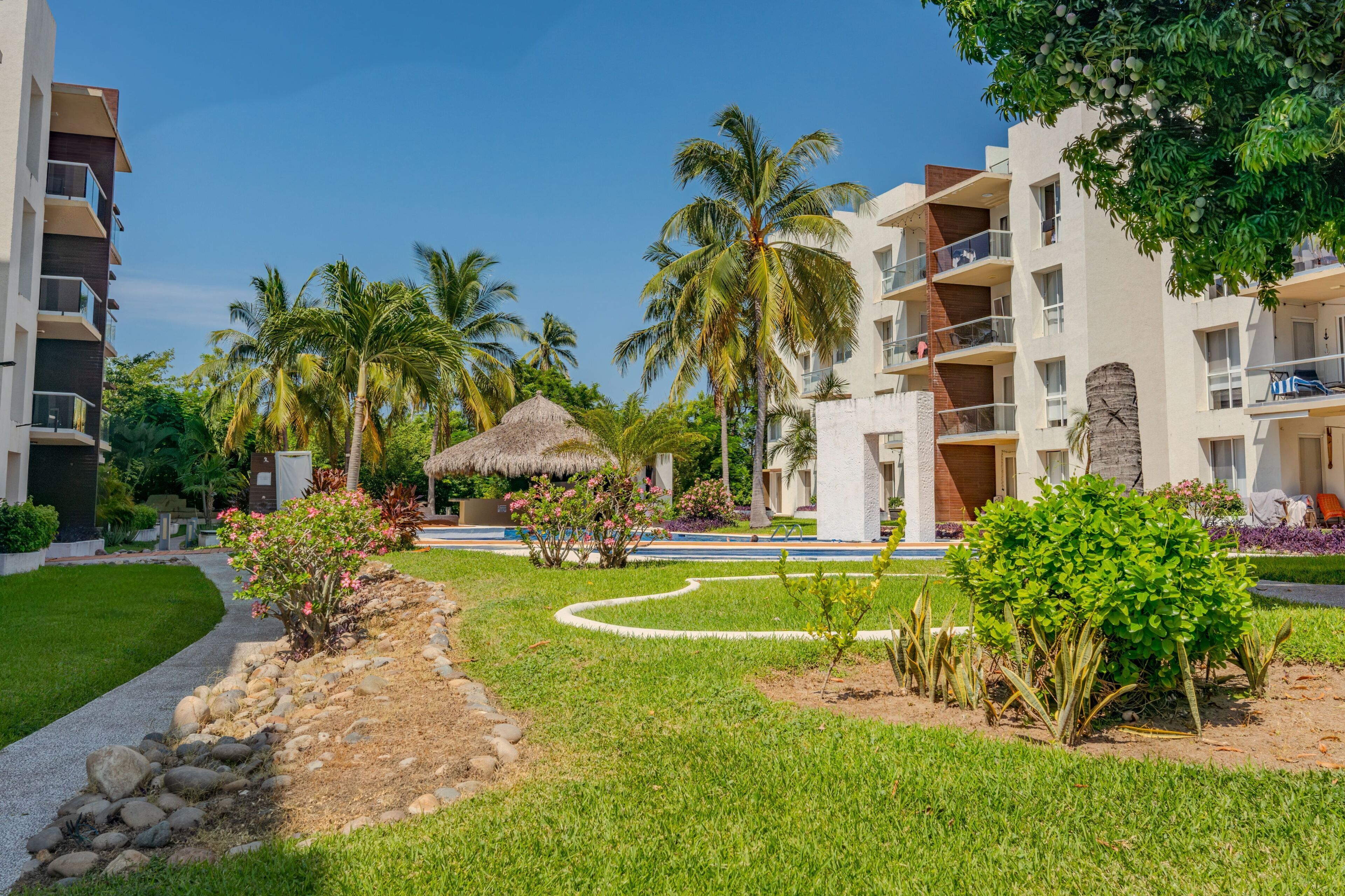 Un resort con edificios de varios pisos, piscina, palmeras y una estructura con techo de paja.