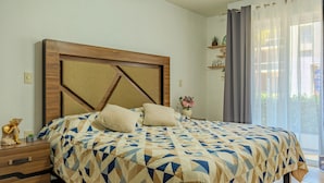 A bedroom with a wooden headboard, a patterned bedspread, a bedside table with a plant, and a window with curtains. - Acapulco Getaway Pool & Jacuzzi 2 BR (Acapulco de Juárez)