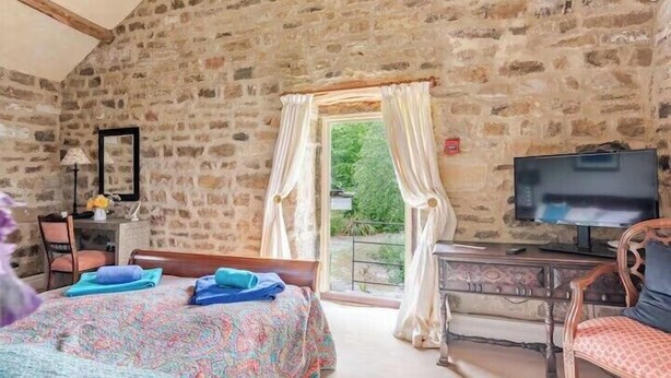 Ein Schlafzimmer mit einer Steinsäule, einem Bett mit gemustertem Überwurf, einem Schreibtisch mit Fernseher und einem Fenster mit Vorhängen.