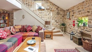 Ein gemütliches Wohnzimmer mit einer Steinwand, einer Treppe, einem Sofa mit gemusterten Kissen, einem Couchtisch und einem Bücherregal.
