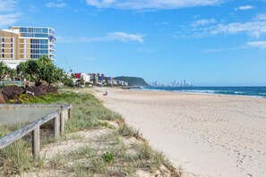 Una playa con una cerca de madera, una zona con césped y un horizonte urbano al fondo.