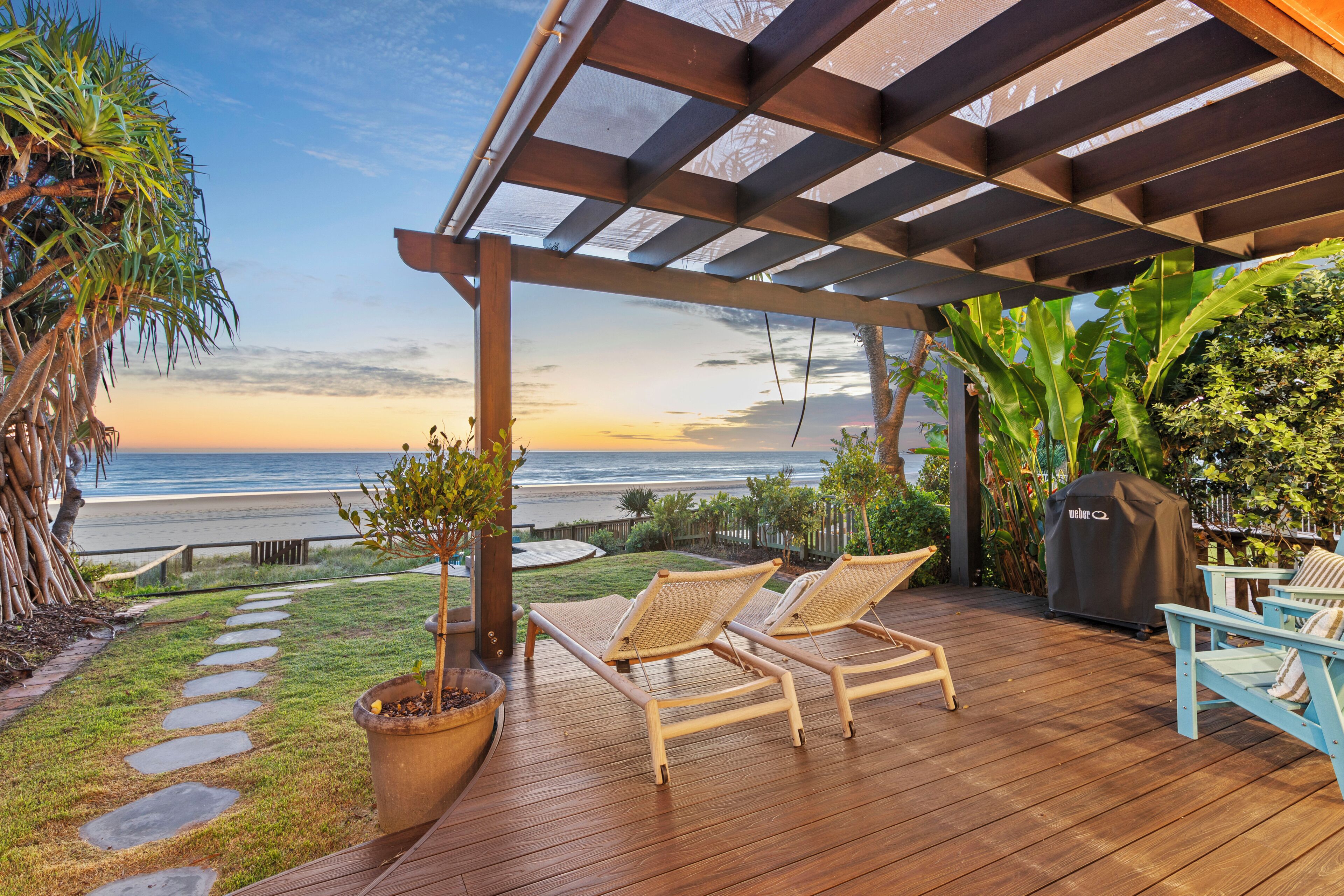 A wooden deck with two wicker chairs and a grill, overlooking a beach with a clear sky.