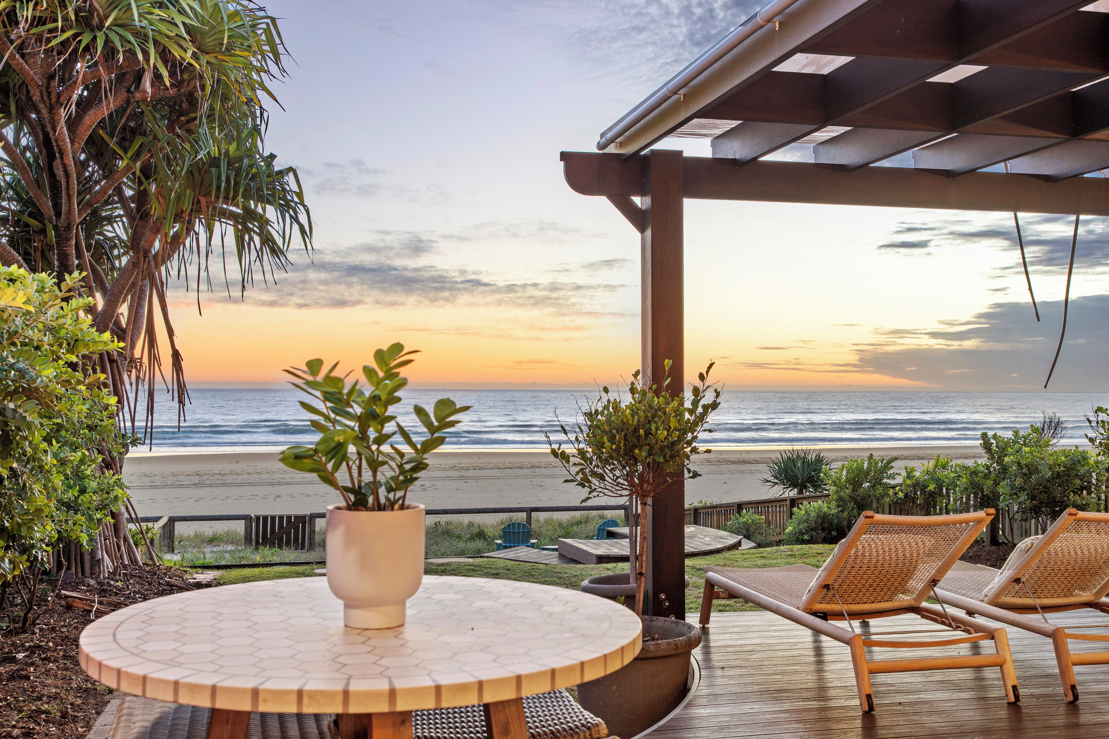 Une terrasse avec une table ronde, deux fauteuils de jardin en osier et une vue sur la plage au coucher du soleil.