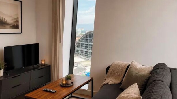 A modern living room with a large window, a flat-screen TV, a dark sofa, and a wooden coffee table. - Salford Serenity - Modern 2 Bedroom Apartment (Salford)