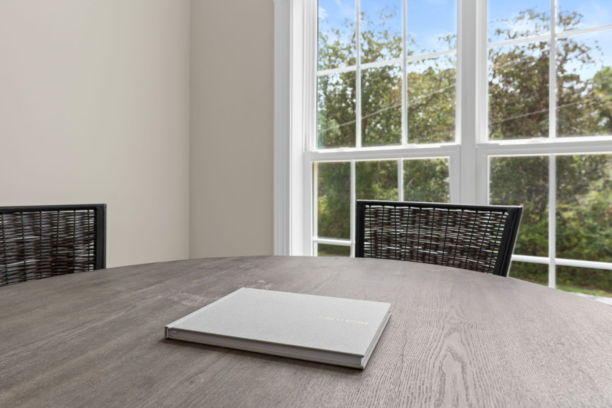 A round table with a grey surface and a book titled 'GUESTROOMS' on it. There are two black chairs with woven backs. The room has white walls and a large window with a view of trees.