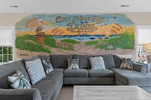 Ein Wohnzimmer mit einer großen Wandmalerei einer Strandlandschaft, einem grauen Ecksofa und Zierkissen.