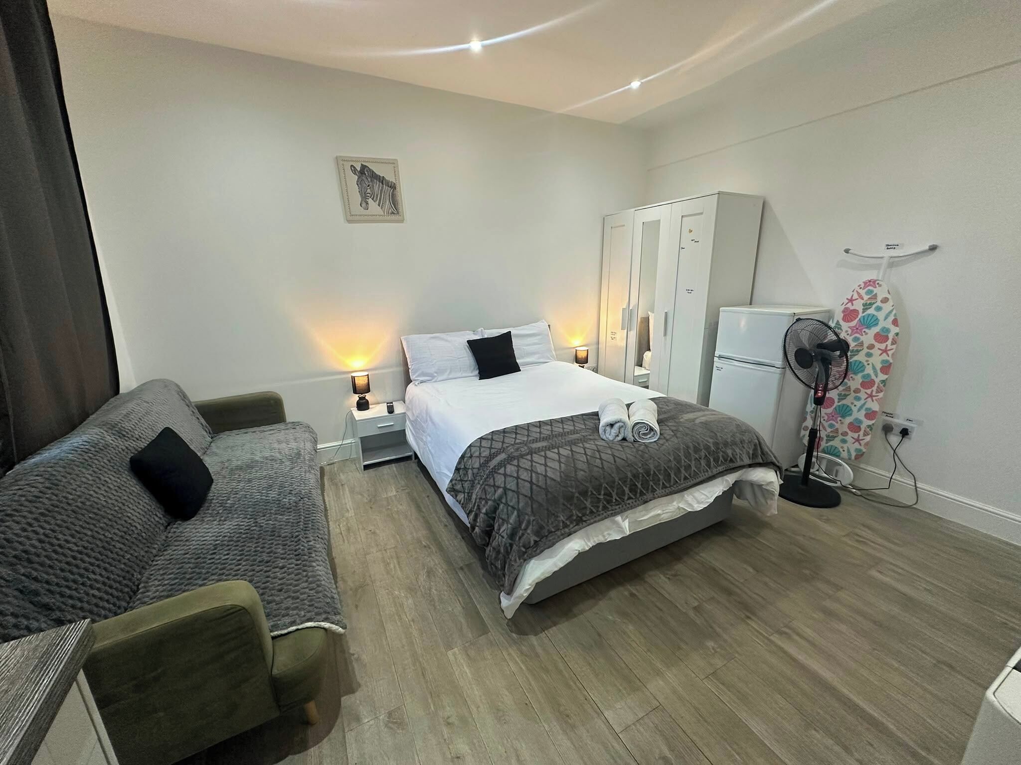 A hotel room with a bed, a sofa, a wardrobe, a desk, a lamp, a fan, and a wall-mounted artwork.