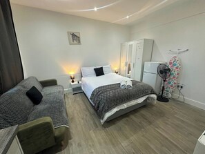 A hotel room with a bed, a sofa, a wardrobe, a desk, a lamp, a fan, and a wall-mounted artwork.