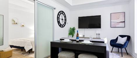 A modern, well-lit room with a bed, a dining area with a table and chairs, a TV mounted on the wall, and a large window.