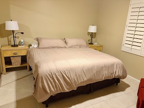 A bedroom with a bed, two bedside tables with lamps, a waste basket, and a window with blinds.