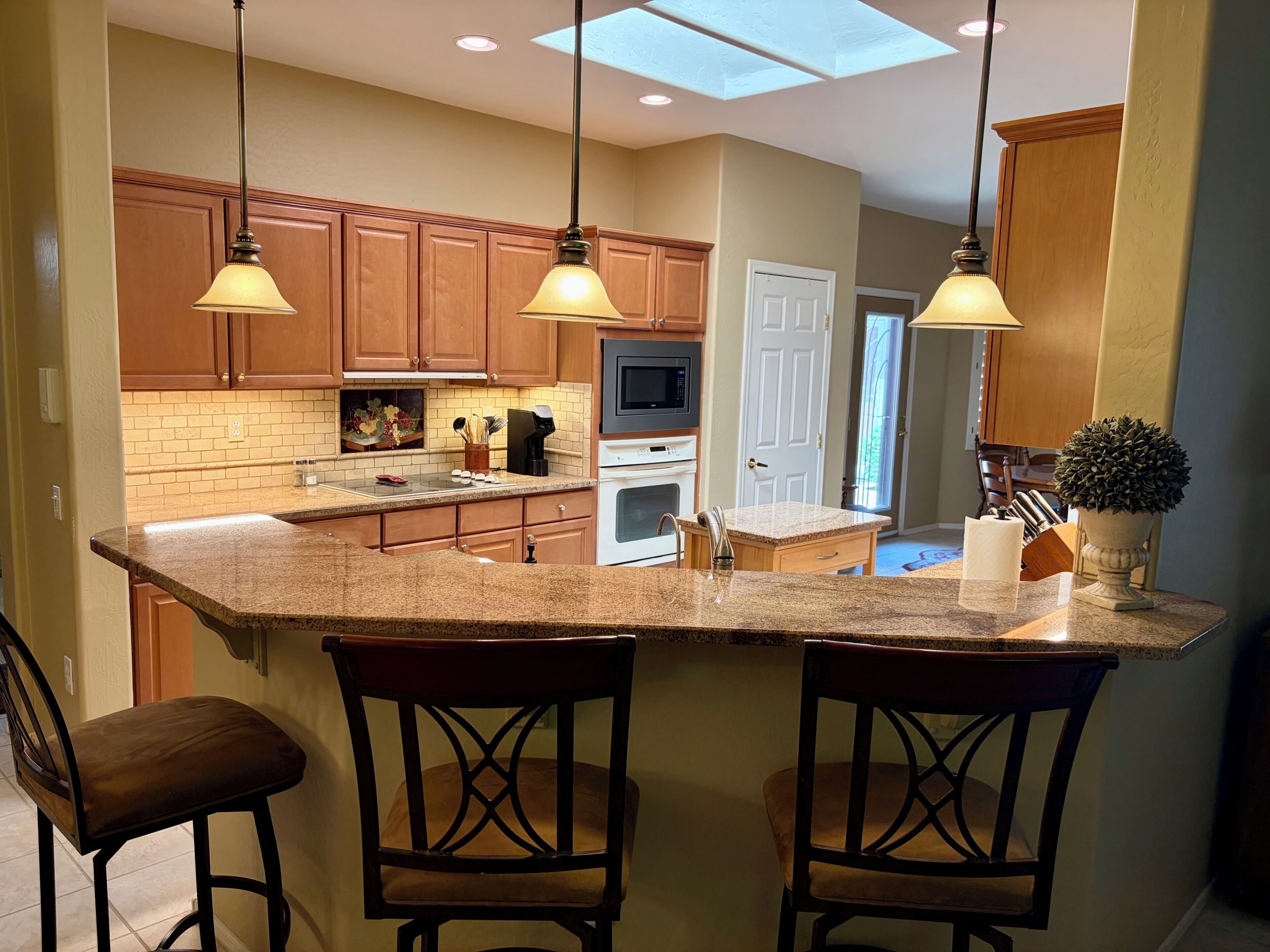A kitchen with a central island, wooden cabinets, a microwave, oven, and a sink. There are two bar stools and pendant lights above the island.