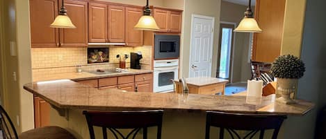 Una cocina con una isla central, armarios de madera, microondas, horno y fregadero. Hay dos taburetes de bar y luces colgantes sobre la isla.