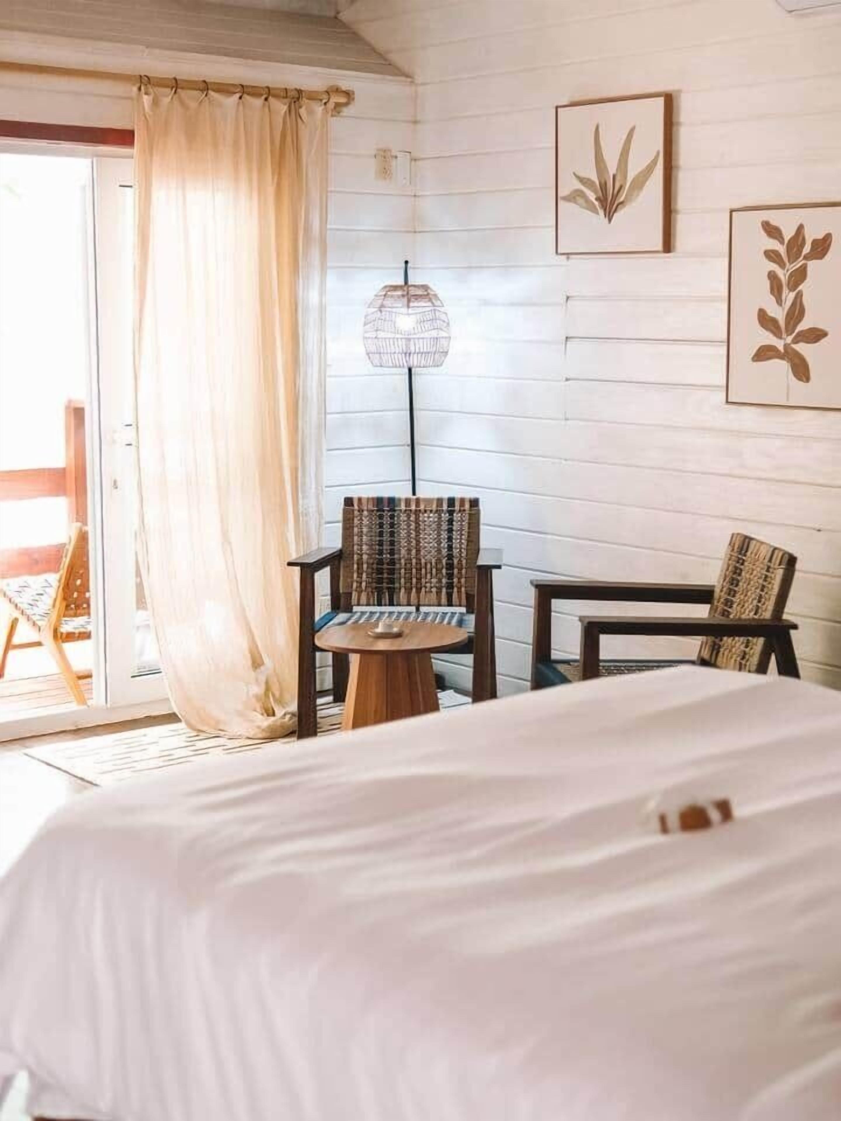 Quarto de hotel com cama, duas cadeiras, uma mesinha, abajur e uma porta de vidro deslizante que dá para uma área externa.