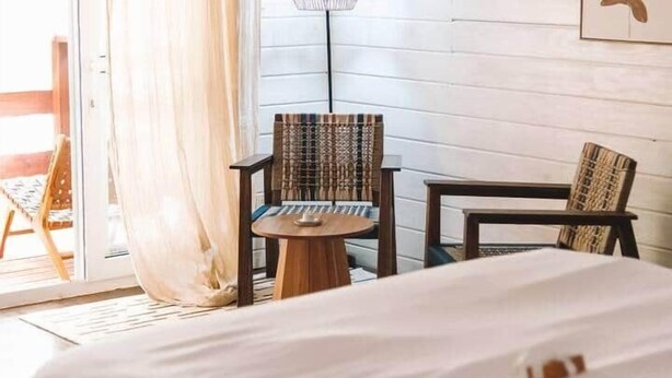 Ett hotellrum med en säng, två stolar, ett litet bord, en lampa och en skjutdörr i glas som leder ut till ett utomhusområde.