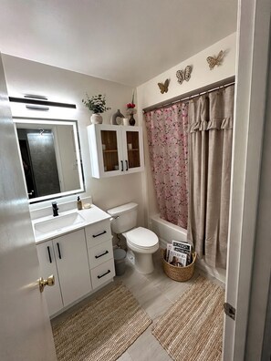 Ein Badezimmer mit einem weißen Waschbecken, einer Toilette, einer Badewanne und einer Duschvorhang. An der Wand sind dekorative Schmetterlinge angebracht und ein Korb mit Zeitschriften steht bereit.