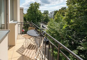Un balcon avec une table et des chaises, donnant sur une rue bordée de voitures garées et d’arbres.