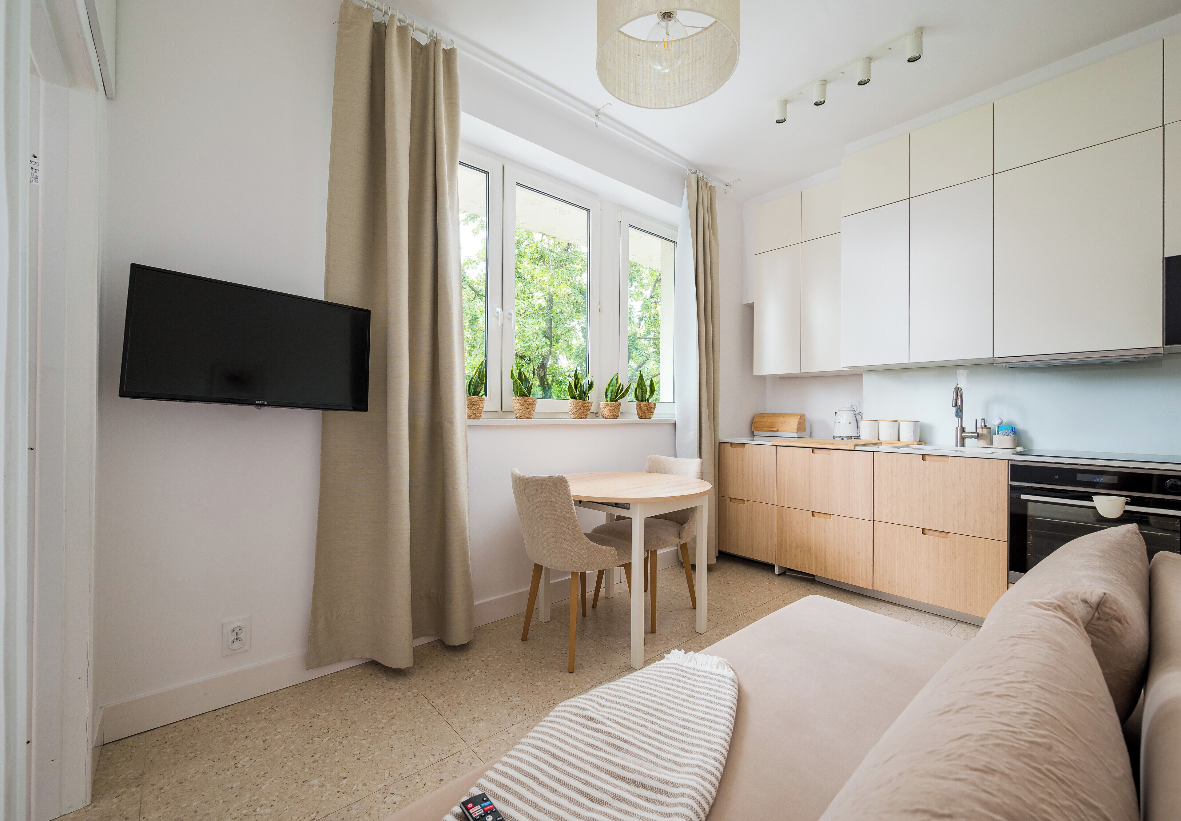 Et moderne, veloplyst rum med et vægmonteret fladskærms-tv, et spisebord med stole, et køkkenområde med skabe og hårde hvidevarer, samt en stor seng med et stribet plaide.