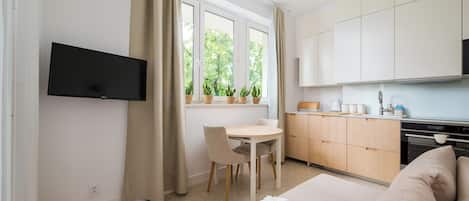Una habitación moderna y luminosa con televisión de pantalla plana en la pared, una mesa de comedor con sillas, una zona de cocina con armarios y electrodomésticos, y una cama grande con una manta a rayas.