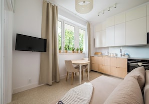 Ett modernt, väl upplyst rum med en platt-TV som är monterad på väggen, ett matbord med stolar, en köksdel med skåp och vitvaror samt en stor säng med ett randigt överkast.