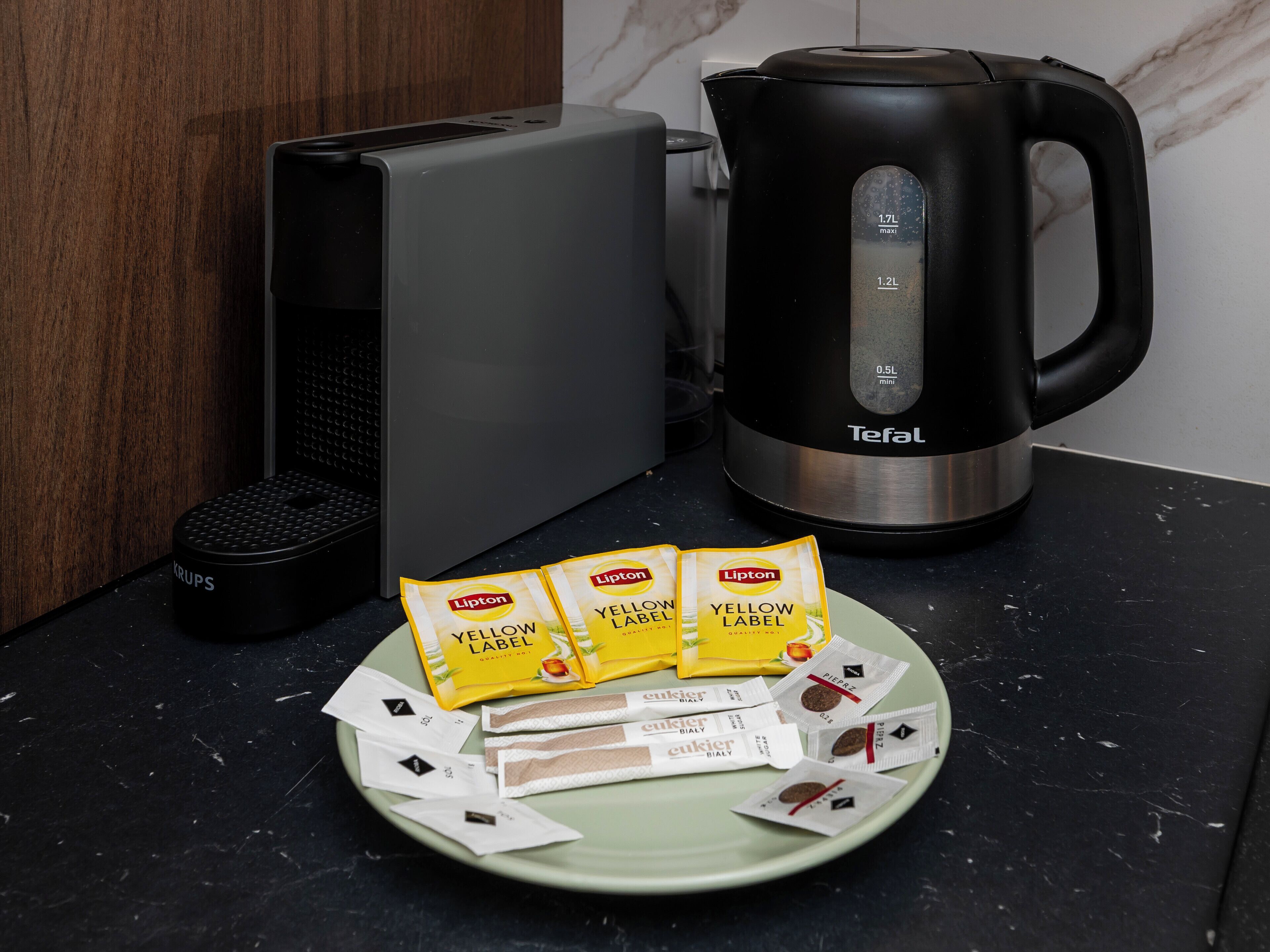 A kitchen area with a coffee maker, a kettle, and a plate with coffee pods.