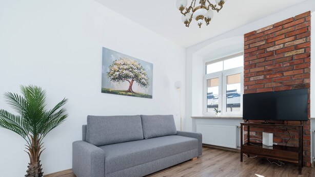 Ett vardagsrum med en grå soffa, en krukväxt, en tavla som föreställer ett träd, en tegelvägg och en tv som står på ett träställ.