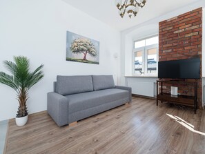 Ein Wohnzimmer mit grauem Sofa, einem Topfpflanzen, einem Baumgemälde, einer Ziegelwand und einem Fernseher auf einem Holzständer.