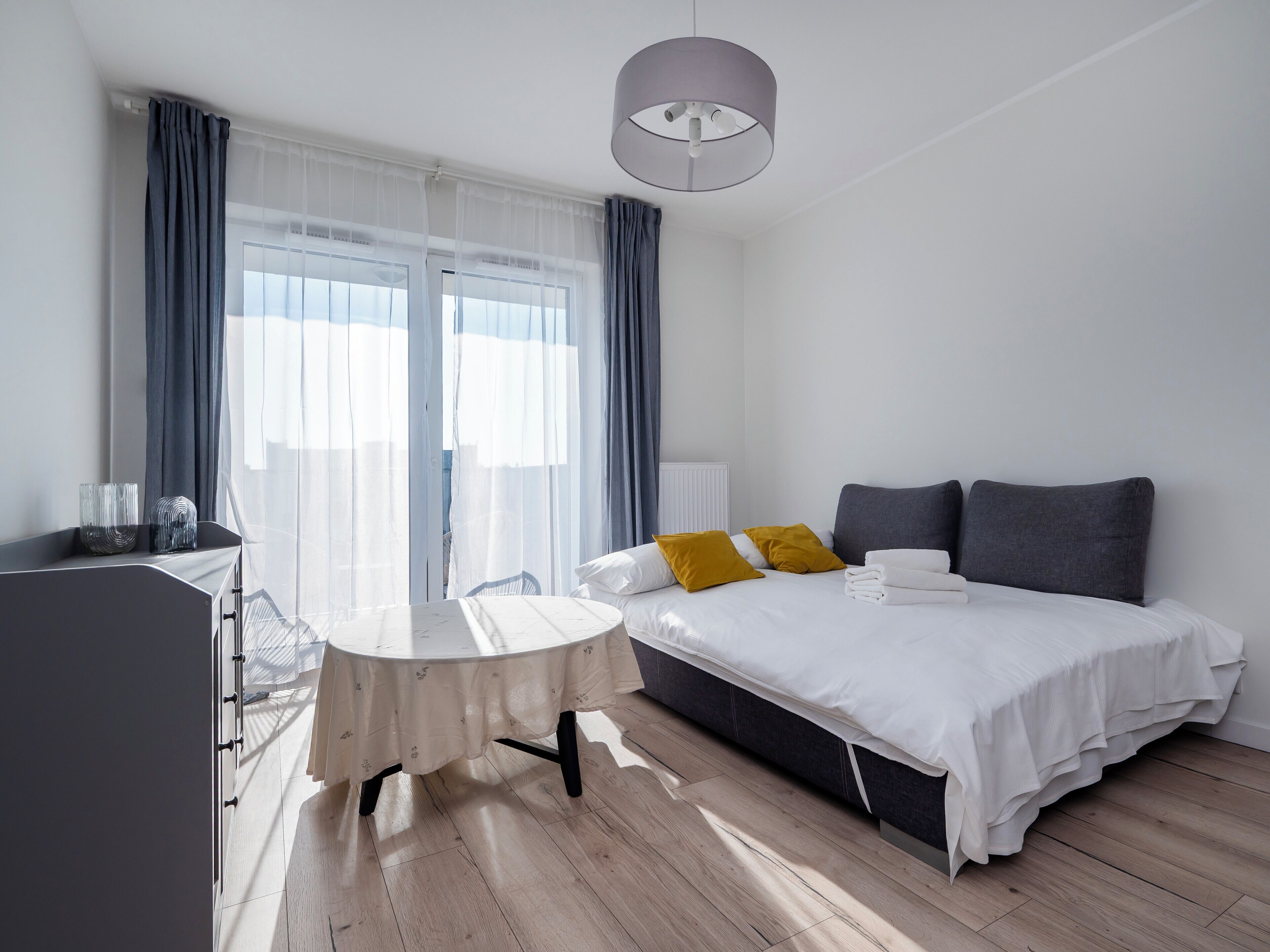 A modern hotel room with a bed, bedside table, and a view of the outdoors through large windows.