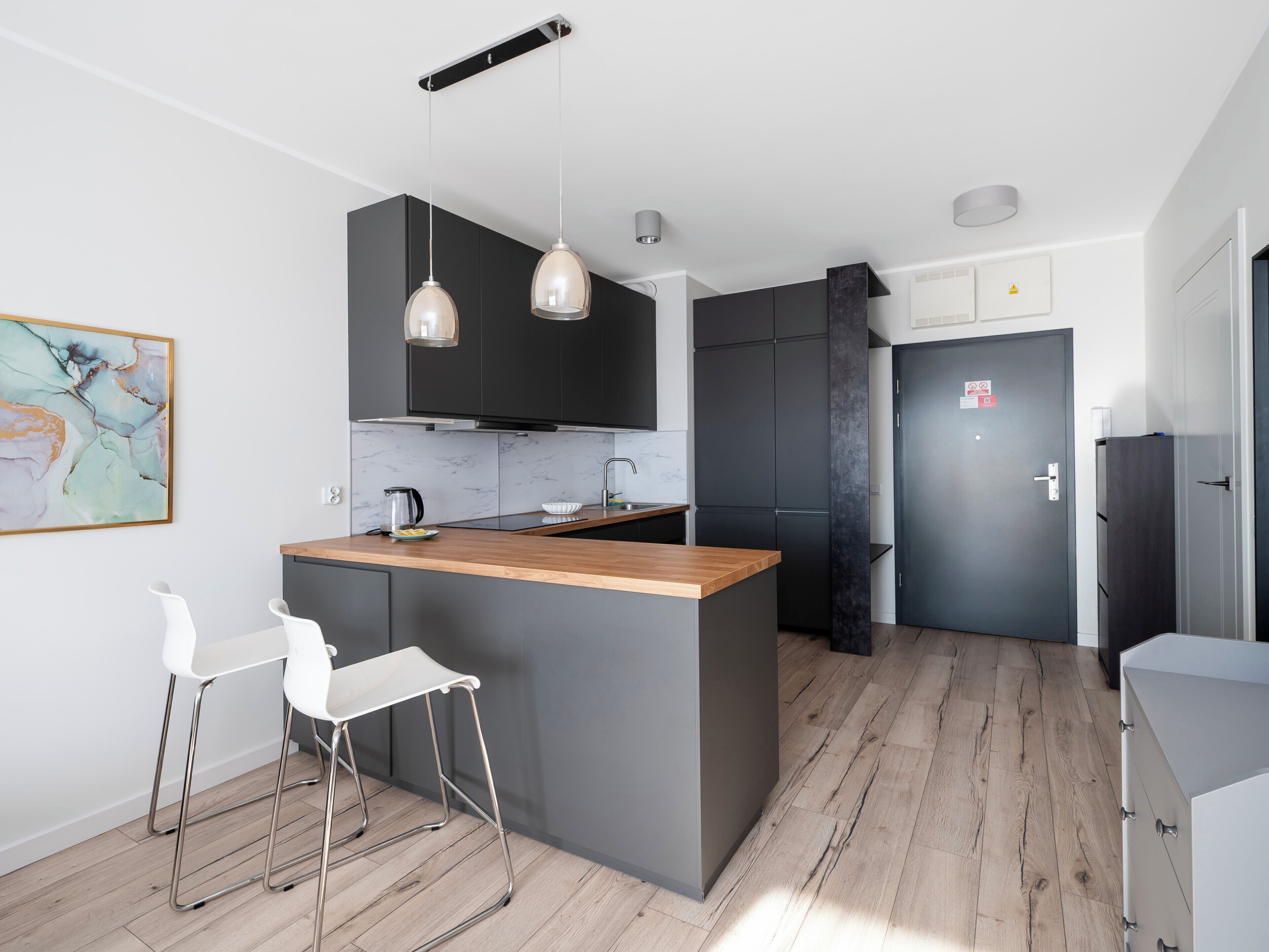 A modern kitchen with a central island, bar stools, pendant lighting, and a dark-colored door.