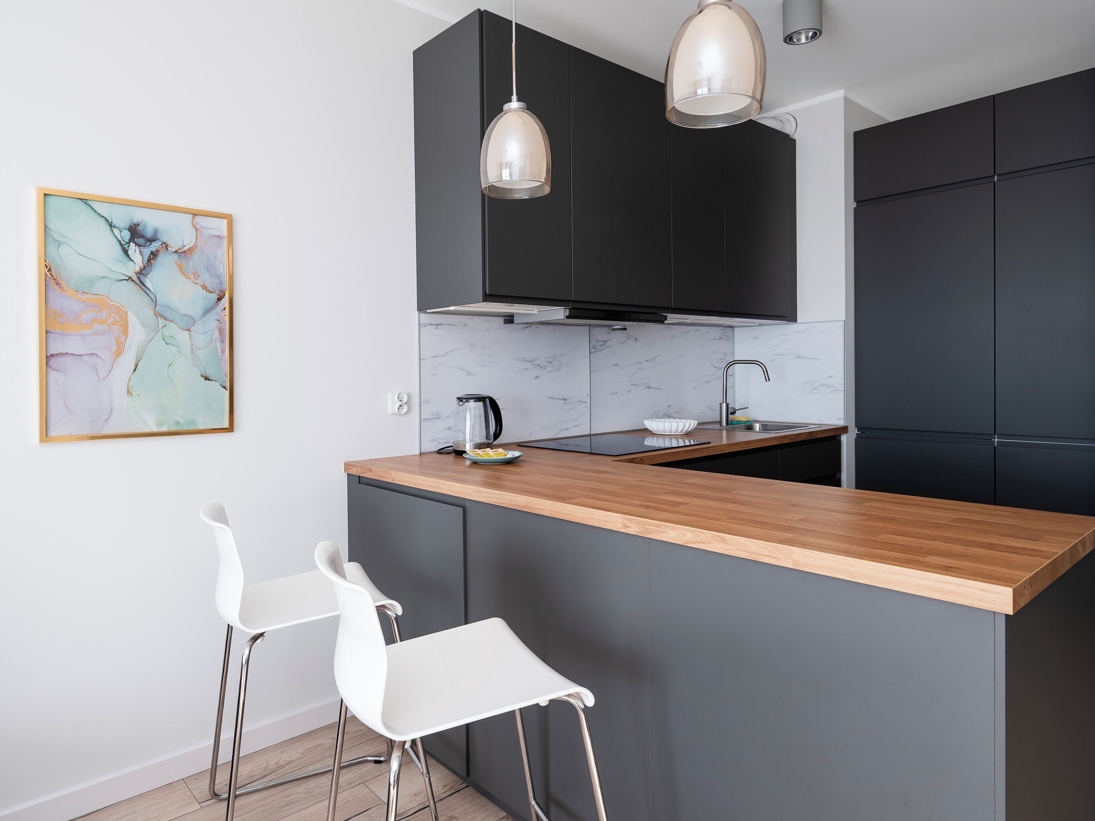 Una cocina moderna con armarios oscuros, encimera de madera, hervidor negro y taburete blanco.