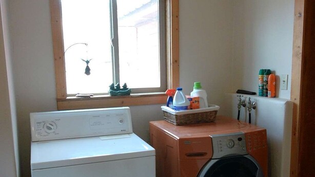 A laundry room with a front-loading washer and dryer, a window with a hanging ornament, and a shelf with cleaning supplies. - Three Bedroom Cabin Escape near Dolores River on Private 300 Acre Ranch! (Dolores)