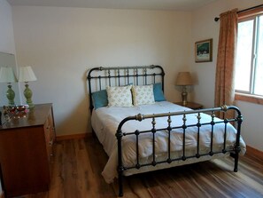 Ein Schlafzimmer mit einem Metallbett, einer Holzkommode, einem Nachttisch mit Lampe und einem fenster mit Vorhängen.