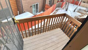 A wooden deck with a metal railing, overlooking a residential area with houses and a red fence.