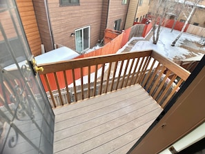 A wooden deck with a metal railing, overlooking a residential area with houses and a red fence.
