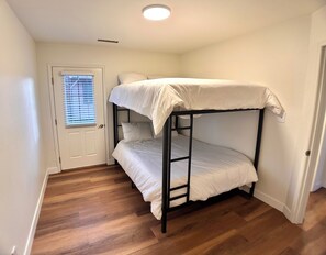 Un lit superposé avec une literie blanche, dans une pièce au plancher en bois et dotée d’une porte avec une fenêtre.