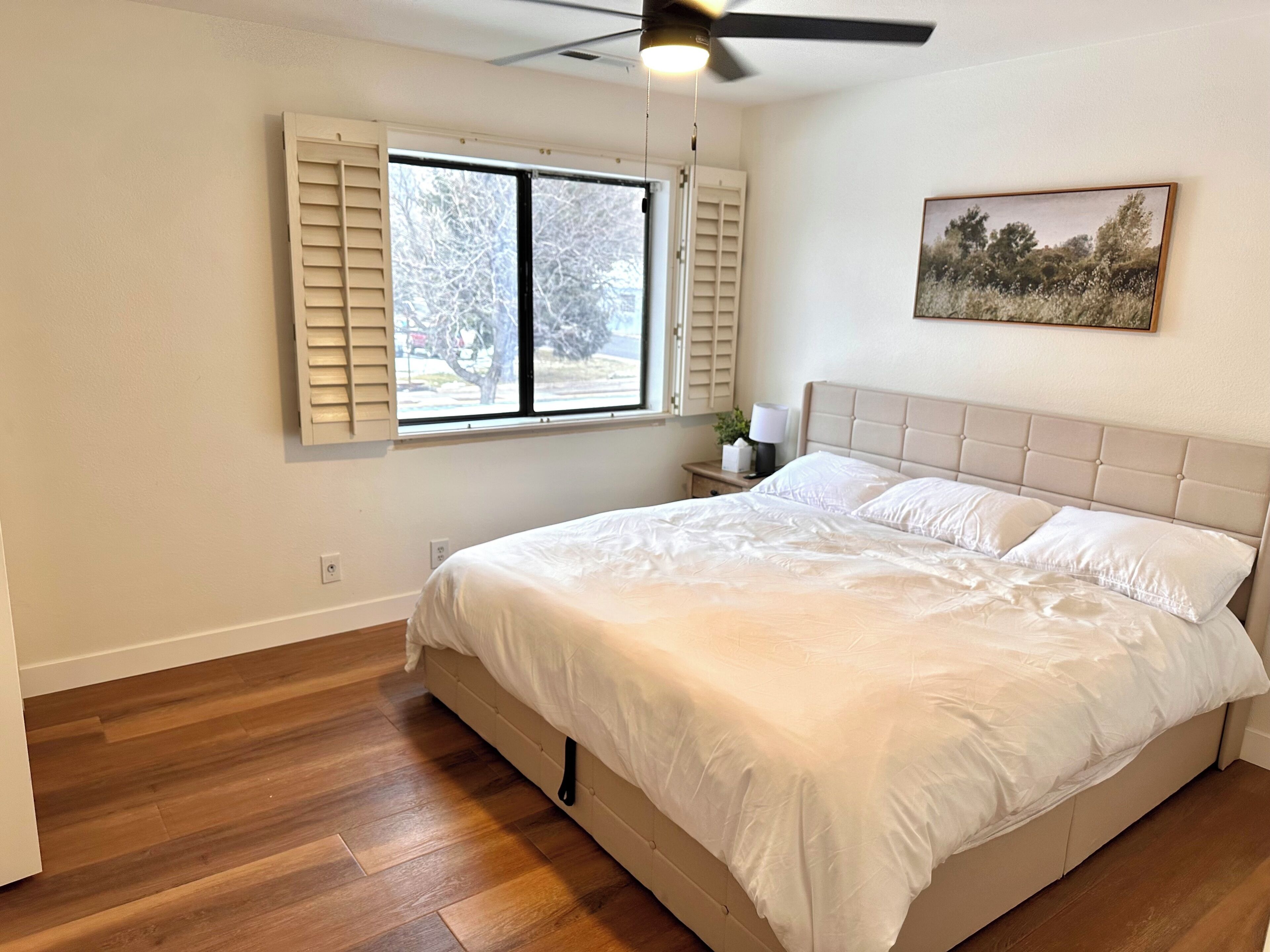 A bedroom with a bed, a window with shutters, a ceiling fan, and a painting on the wall.