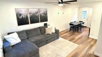 A living room with a gray sectional sofa, a white rug, a dining table with chairs, and a ceiling fan.