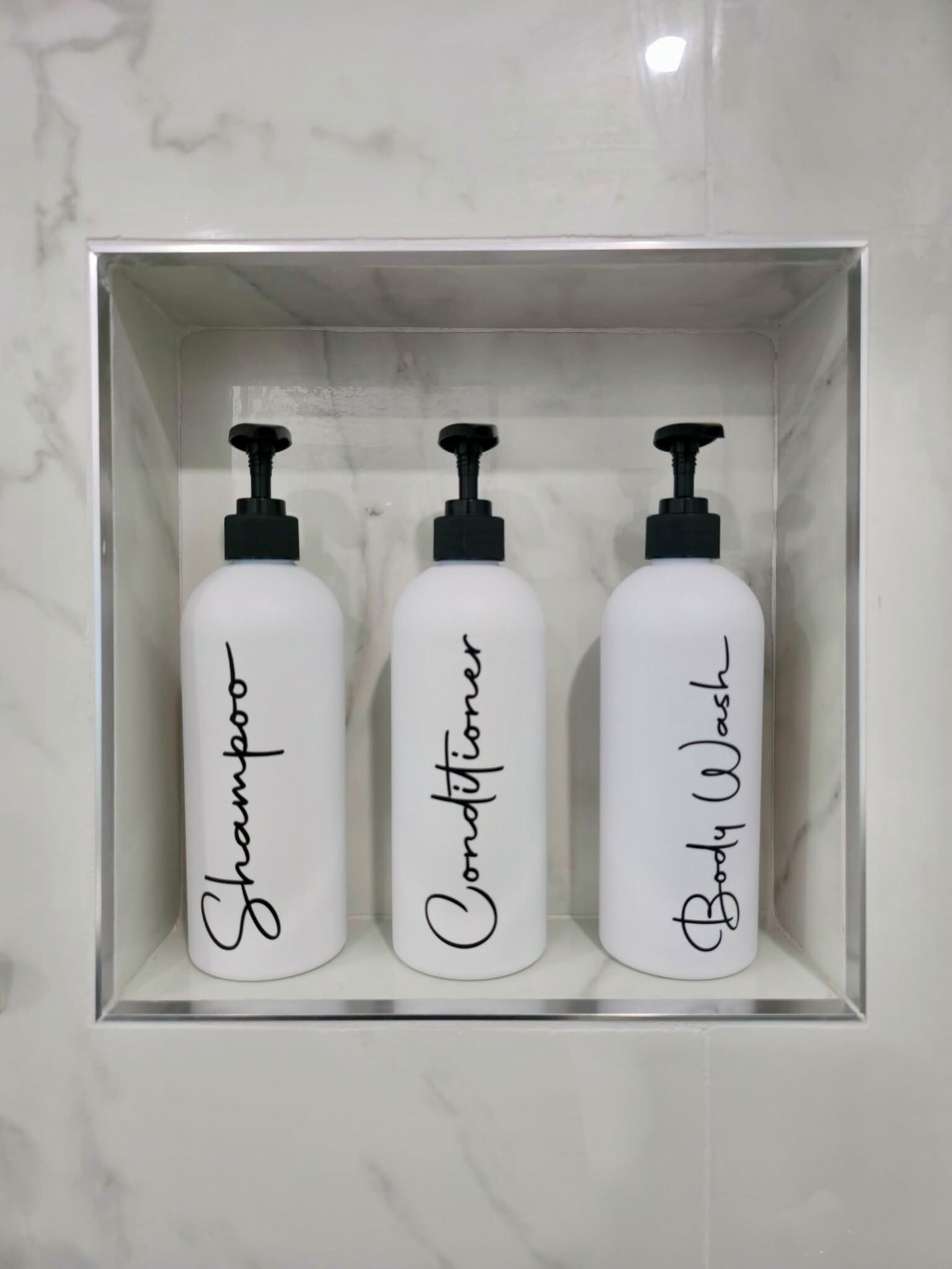 Drei weiße Flaschen mit schwarzen Dosierern, beschriftet mit 'Shampoo', 'Conditioner' und 'Body Wash', in einer weißen Schachtel.