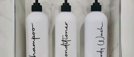 Trois flacons blancs avec des pompes noires, étiquetés « Shampoing », « Après-shampoing » et « Gel douche », dans une boîte blanche.