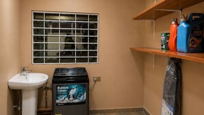 A bathroom with a sink, a window with bars, a heater, and shelves with cleaning supplies.