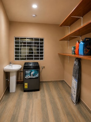 A bathroom with a sink, a window with bars, a heater, and shelves with cleaning supplies.