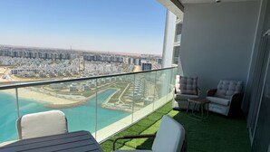 A balcony with a view of a coastal cityscape, a pool, and artificial grass. - Luxury 1-bedroom in Gate tower with double View (New Alamein)