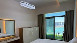 A hotel room with a bed, a bedside table, a mirror, a trash bin, and a large window with a view of a body of water and buildings. - Luxury 1-bedroom in Gate tower with double View (New Alamein)