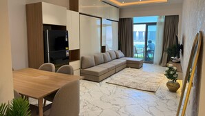 A modern living room with a sectional sofa, a dining table, and a kitchen area with a refrigerator and cabinets. - Luxury 1-bedroom in Gate tower with double View (New Alamein)