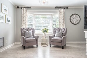 A room with two armchairs facing a bay window, a round wall clock, and framed pictures on the wall. - The Mad Hatter Inn - In the Village of Pinehurst! (Pinehurst)