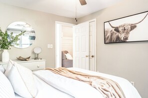 A bedroom with a bed, a bedside table with a mirror, a wall-mounted picture, and a door leading to another room. - The Mad Hatter Inn - In the Village of Pinehurst! (Pinehurst)
